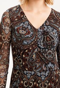 Nahaufnahme des Kleides einer Frau mit V-Ausschnitt und filigranen Paisleymustern in kräftigen Farben, darunter Braun, Blau und Rot.