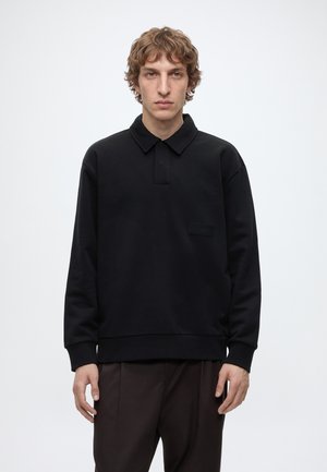 Jeune homme aux cheveux ondulés châtain clair, portant un polo noir à manches longues et un pantalon foncé, debout devant un fond blanc uni.