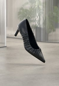 Black leather high-heel pump with a pointed toe, featuring gathered texture along the opening and a mid-height stiletto heel.