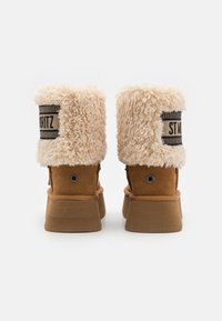 Stivaletti alla caviglia in suede marrone con rivestimento soffice color crema, caratterizzati da una fascia lavorata con l'etichetta "ST. MORITZ" e un accento circolare sul tallone.