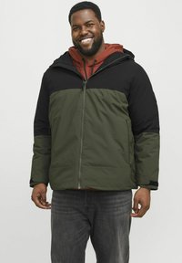 Groene en zwarte geïsoleerde jas met rits aan de voorkant, capuchon en zijzakken; gedragen over een rode hoodie; heeft een gestructureerde stof.