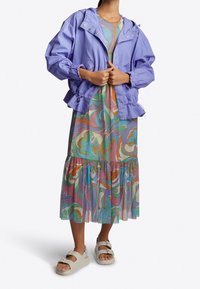 Paarse lichtgewicht jas met elastische manchetten, gedragen over een vloeiende, veelkleurige jurk met zwirls en een gerimpelde zoom. Beigekleurige sandalen maken de look compleet.