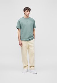 Jaunas vyras stovintis, dėvintis žalią „Nike“ marškinėlį, šviesių spalvų kelnes ir baltus sportbačius, priešais paprastą šviesiai pilką foną.