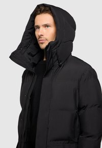 Giubbotto puffer nero con un ampio cappuccio, chiusura con zip frontale e tasche laterali. Presenta una texture trapuntata e cordini regolabili.