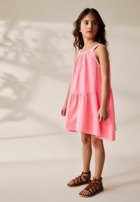 Vestido sin mangas de color rosa con escote fruncido y dobladillo en capas, hecho de una tela ligera; combinado con sandalias marrones con tiras.