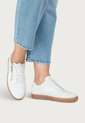 Zwei Füße mit weißen Leder-Schnürschuhen und hellbraunen Sohlen, kombiniert mit hellblauen, gekürzten Jeans vor einem einfarbigen Hintergrund.