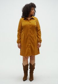 Sennepsfarget kordfløyelskjortekjole med lange ermer, knapper foran og en litt innsvingt nederkant. Kombinert med brune knelange støvler.