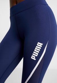 Námořnické modré sportovní legíny vyrobené z pružné látky, s bílým logem PUMA a diagonálním bílým akcentem poblíž bočního švu.