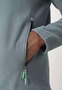 Fleece jopica v mehki sivi barvi z zadrgo in žepom. Ima gladko teksturo, standardne manšete in zeleno oznako pritrjeno na zadrgo.