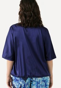 Blusa di seta navy con maniche corte, texture liscia e vestibilità ampia. Abbinata a pantaloni con motivo floreale nei toni del blu e del bianco.
