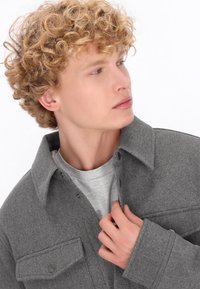 Jachetă din flanel gri, cu guler ascuțit, nasturi cu capsuță și un buzunar la piept, suprapusă peste un tricou gri deschis cu guler rotund.