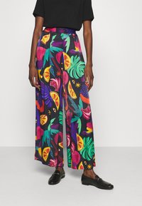 Une personne portant un pantalon à jambes larges avec des motifs colorés de fruits tropicaux et de feuilles, associé à des mocassins noirs et un haut noir uni.