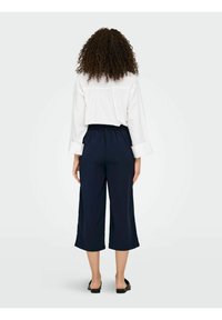 Pantalon court marine à taille élastique, associé à une chemise blanche boutonnée avec un dos plissé, sur un fond neutre.
