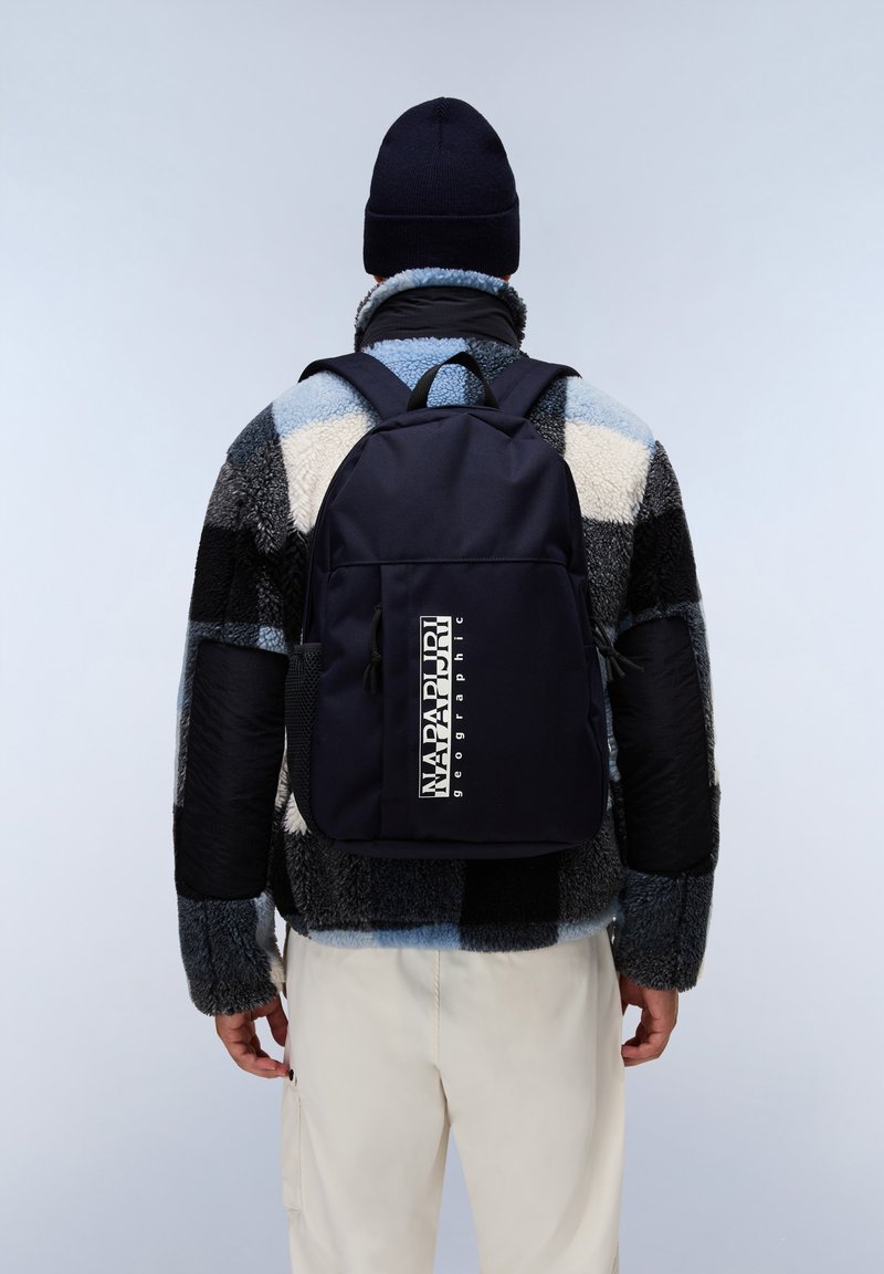 Zaino blu navy con un unico scomparto, caratterizzato da un grande logo e una tasca laterale in rete, abbinato a una giacca in pile multicolore e testurizzata.