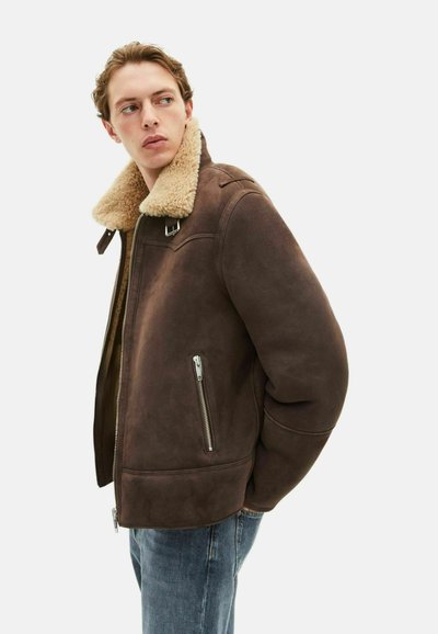 Chaqueta de shearling marrón con un acolchado collar en tono beige, bolsillos laterales con cremallera y un ajuste ligeramente holgado. Textura de ante suave y diseño estructurado.