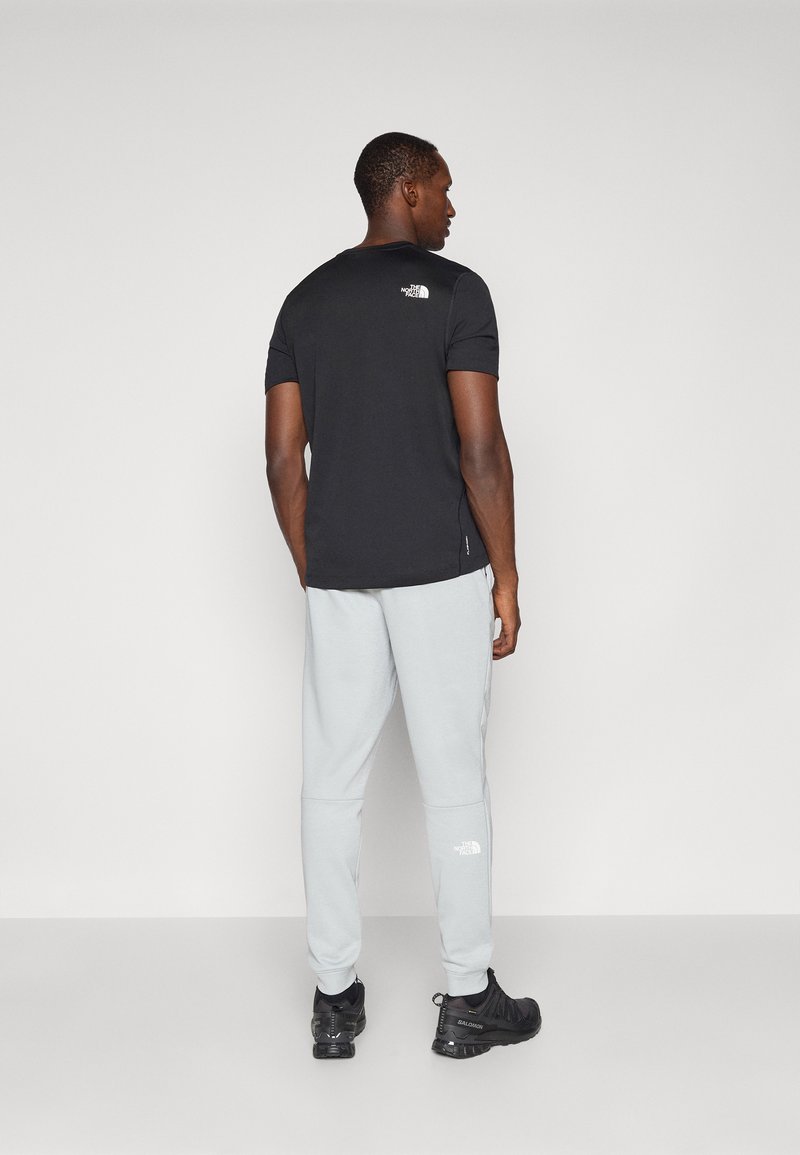 T-shirt noir à manches courtes avec un petit logo blanc dans le haut du dos, associé à un pantalon de jogging gris clair et des chaussures noires.