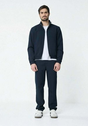 Veste de survêtement marine avec fermeture éclair blanche, assortie à un pantalon. Le modèle porte un t-shirt blanc et des chaussures de sport grises avec des accents blancs.
