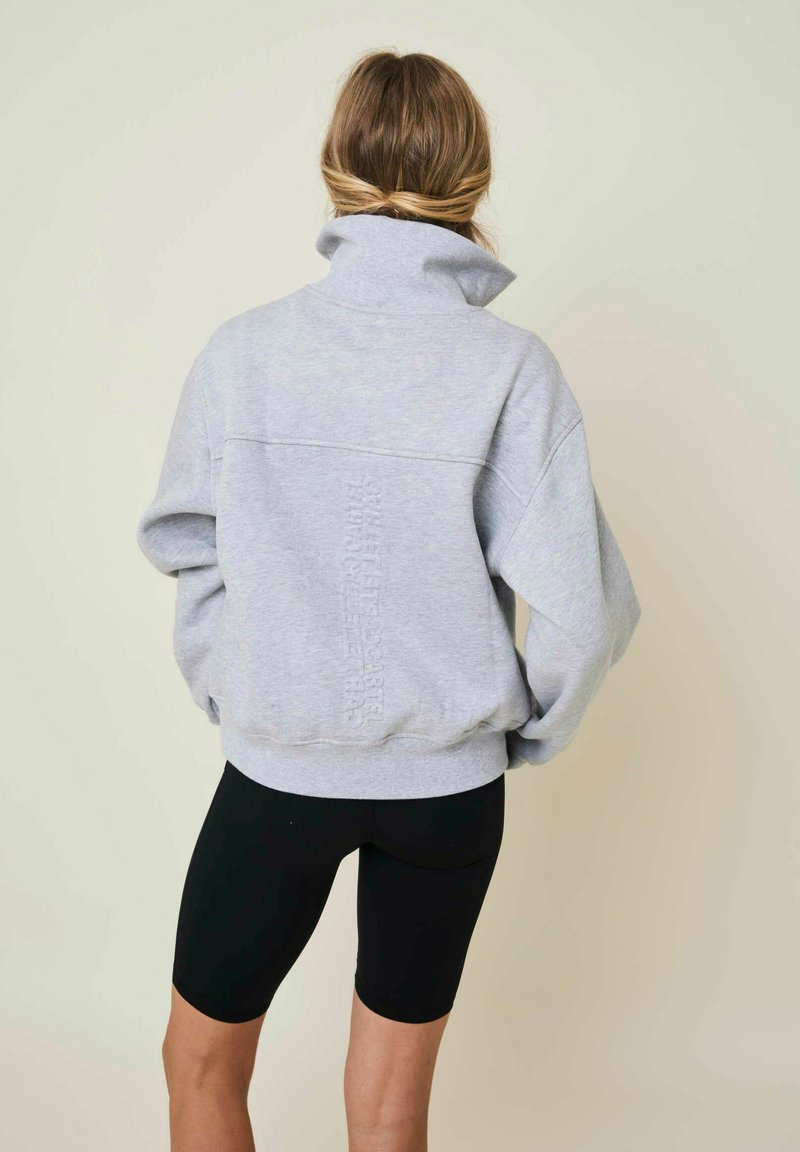 Άτομο με μαλλιά δεμένα πίσω φοράει ανοιχτό γκρι φούτερ με ψηλό γιακά και μαύρα ποδηλατικά σορτς, όρθιο σε απλό φωτεινό φόντο.
