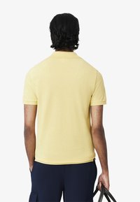 Polo giallo con maniche corte e colletto classico, caratterizzato da una texture liscia. La persona sta tenendo una borsa nera.