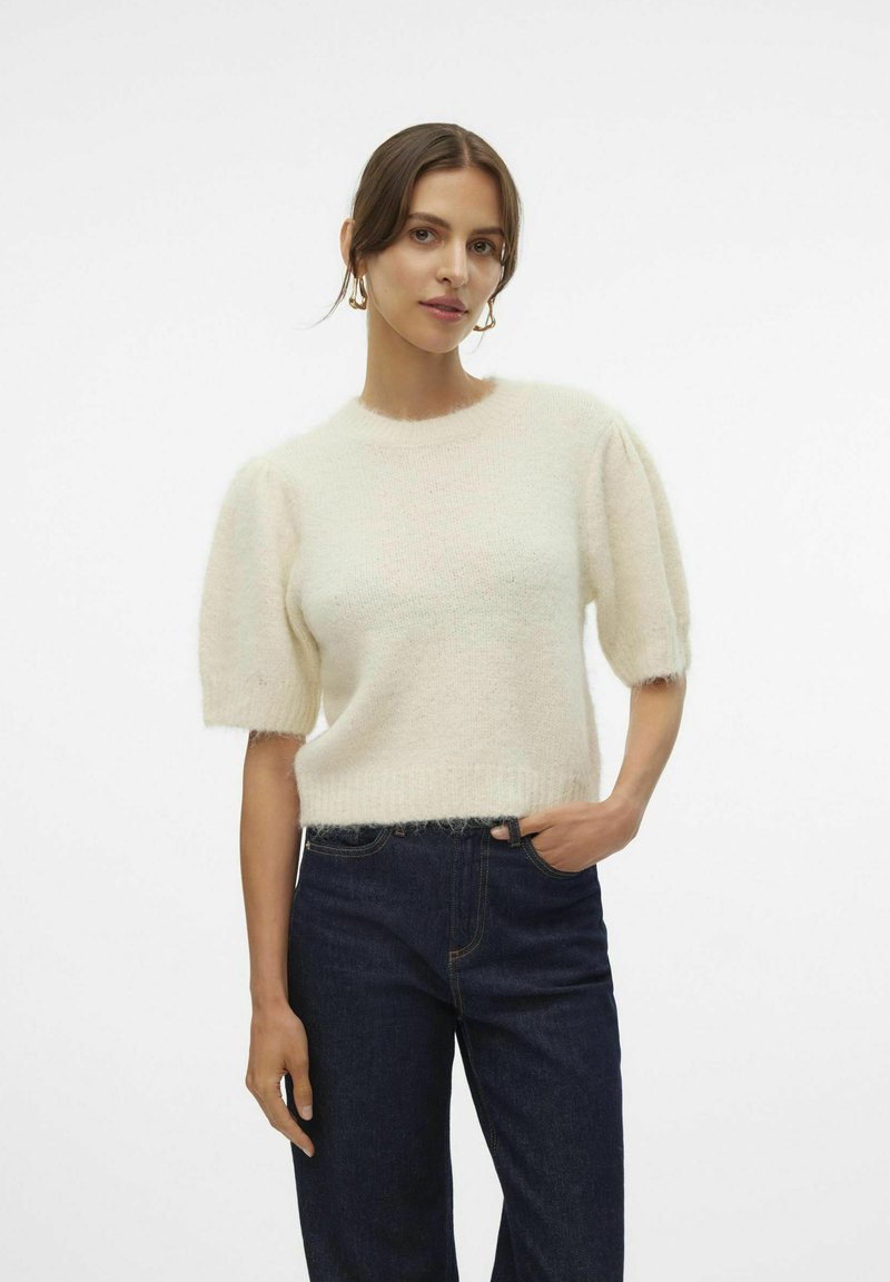 Lichtcrème korteke mouwen trui met een zachte, fuzzy textuur en een ribbelrand. Gecombineerd met donkerblauwe jeans, weergegeven tegen een effen achtergrond.