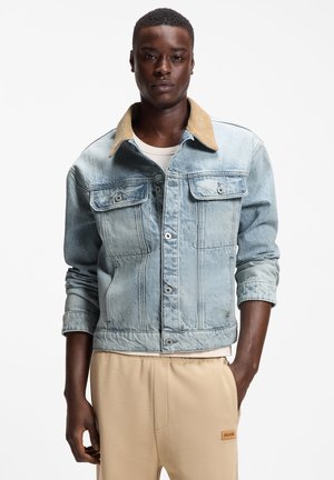 Ung mand iført en lyseblå denimjakke med en tanfarvet krave og beige bukser, stående foran en ensartet hvid baggrund.