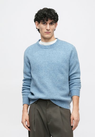 Gaiši zils adīts džemperis ar apaļu kakla izgriezumu un ribotiem piedurknēm. Audums izskatās mīksts; uzvilkts virs baltas kaites un saskaņots ar tumšiem biksēm.