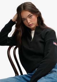Zwarte gebreide trui met een hoge kraag en een halve ritssluiting, voorzien van geribbelde manchetten. Draagt over een witte shirt, gecombineerd met lichtblauwe denim.