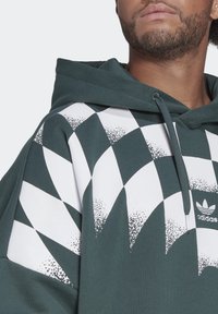 Man i mörkgrön Adidas-hoodie med vit och spräcklig rutmönster över bröstet och ärmarna, med snoddar på huvan.