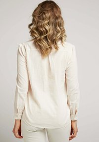 Blouse à manches longues de couleur beige clair en tissu texturé, dotée de poignets boutonnés et d'un ourlet arrondi, avec un design dos uni.