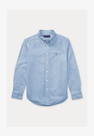 Lys blå skjorte med lange ermer, krage og hvite knapper. Har en liten brodert logo på brystet. Jevn tekstur.