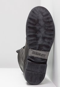 Podeszwa czarnego buta ustawiona pod kątem, ukazująca głęboki wzór bieżnika oraz nazwę marki "REPLAY" wytłoczoną w części pięty.