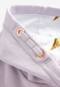 Lavendelfarvet tekstil med en glat tekstur, der har en rosegold øsken og et vævet mærkat, som delvist viser et blomsterdesign nedenunder.