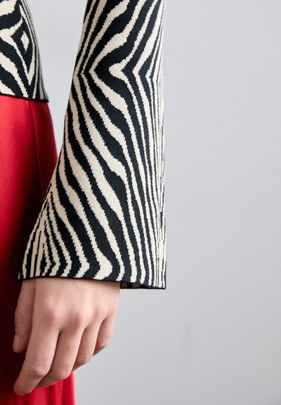 Μαύρη και λευκή ριγέ μανσέτα με υφή, σε συνδυασμό με ένα λείο κόκκινο ύφασμα από κάτω. Κοντινή λήψη ενός χεριού που στηρίζεται σε ύφασμα.