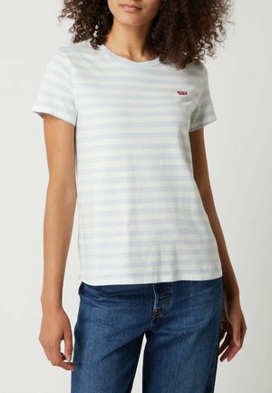 Femme portant un t-shirt à manches courtes rayé bleu clair et blanc et un jean bleu, debout avec les bras relâchés le long du corps.