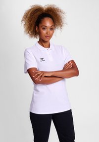 Witte polo met korte mouwen, voorzien van een zwart logo op de linkerborst. Soepele stof en met kraag ontwerp. Donkere broek niet gedetailleerd.