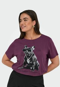 Lilla T-shirt med korte ærmer, der har en grafik af en fransk bulldog iført solbriller og halskæde, med tekst trykt under billedet.