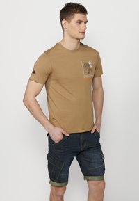 T-shirt marron à manches courtes avec un imprimé graphique et du texte sur le devant, associé à un short cargo en denim foncé avec des ourlets roulés.