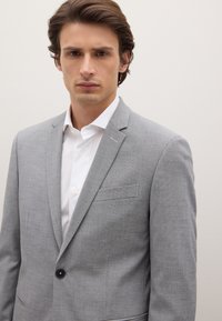 Lichtgrijs colbert met een gestructureerde afwerking, een enkele knoopsluiting en een borstzak, gecombineerd met een wit overhemd met kraag eronder.