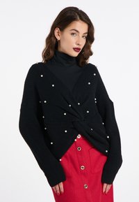 Pullover nero a maglia con dettaglio frontale torcigliato, decorato con perle bianche, abbinato a una gonna rossa con bottoni e tasche.