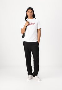 Hvit bomulls t-skjorte med rød grafisk tekst, kombinert med svarte bukser. Modellen har på seg hvite sneakers og holder en liten svart veske.