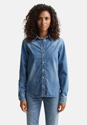 Frau mit lockigem Haar, die ein mittelblaues Jeanshemd mit Knöpfen und passende Jeans trägt, steht vor einem schlichten weißen Hintergrund.