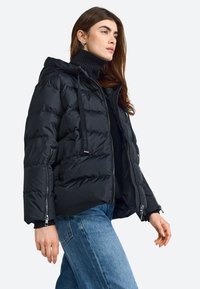 Zwarte pufferjas met een capuchon, voorzien van elastische manchetten, zijritsen en een strakke afwerking. Ggedragen over een zwarte coltrui en blauwe spijkerbroek.