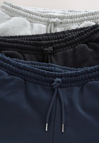 Trzy złożone spodnie dresowe w kolorach szarym, czarnym i granatowym, ułożone w stos z widocznymi elastycznymi pasami i zawiązanymi troczkami.