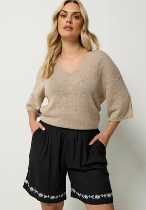Vrouw met lang blond haar die een beige V-hals trui en zwarte shorts met witte bloemenborduurwerk draagt, handen in de zakken, staand tegen een effen achtergrond.