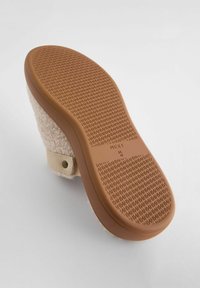 Chaussure beige avec un dessus en laine douce et texturée, et une semelle en caoutchouc avec un motif géométrique pour l'adhérence. Sangle réglable avec un bouton-pression en métal.