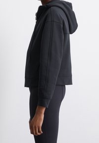 Svart kort luvtröja med dragkedja, sidofickor och ribbade muddar. Mjuk textur och minimalistisk design.