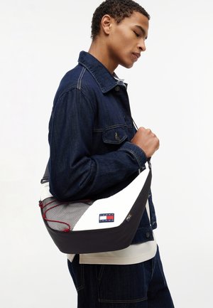 Junger Mann in dunkler Jeansjacke trägt eine schwarz-weiße Umhängetasche mit Netzfach und Tommy Jeans-Logo.