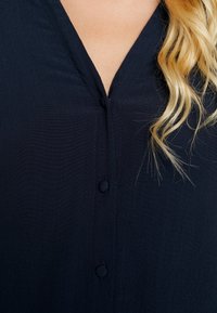 Gros plan sur un chemisier noir à boutons avec un col en V, montrant une partie de cheveux blonds ondulés sur le côté droit.