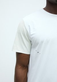 Tricou sportiv alb cu mânecă scurtă, cu o textură subtilă, având o mică gaură în partea inferioară din față, conceput pentru confort și mobilitate.