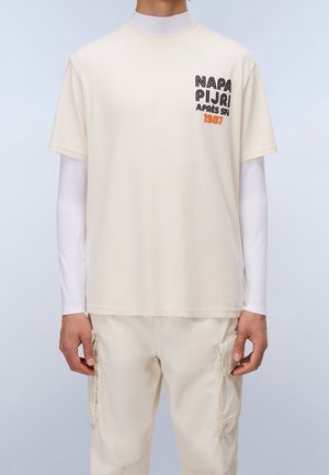 Person iført en cremefarvet kortærmet t-shirt med sort og orange "Napapijri Après Ski 1987" tekst, lagdelt over en hvid langærmet top og cremebukser.